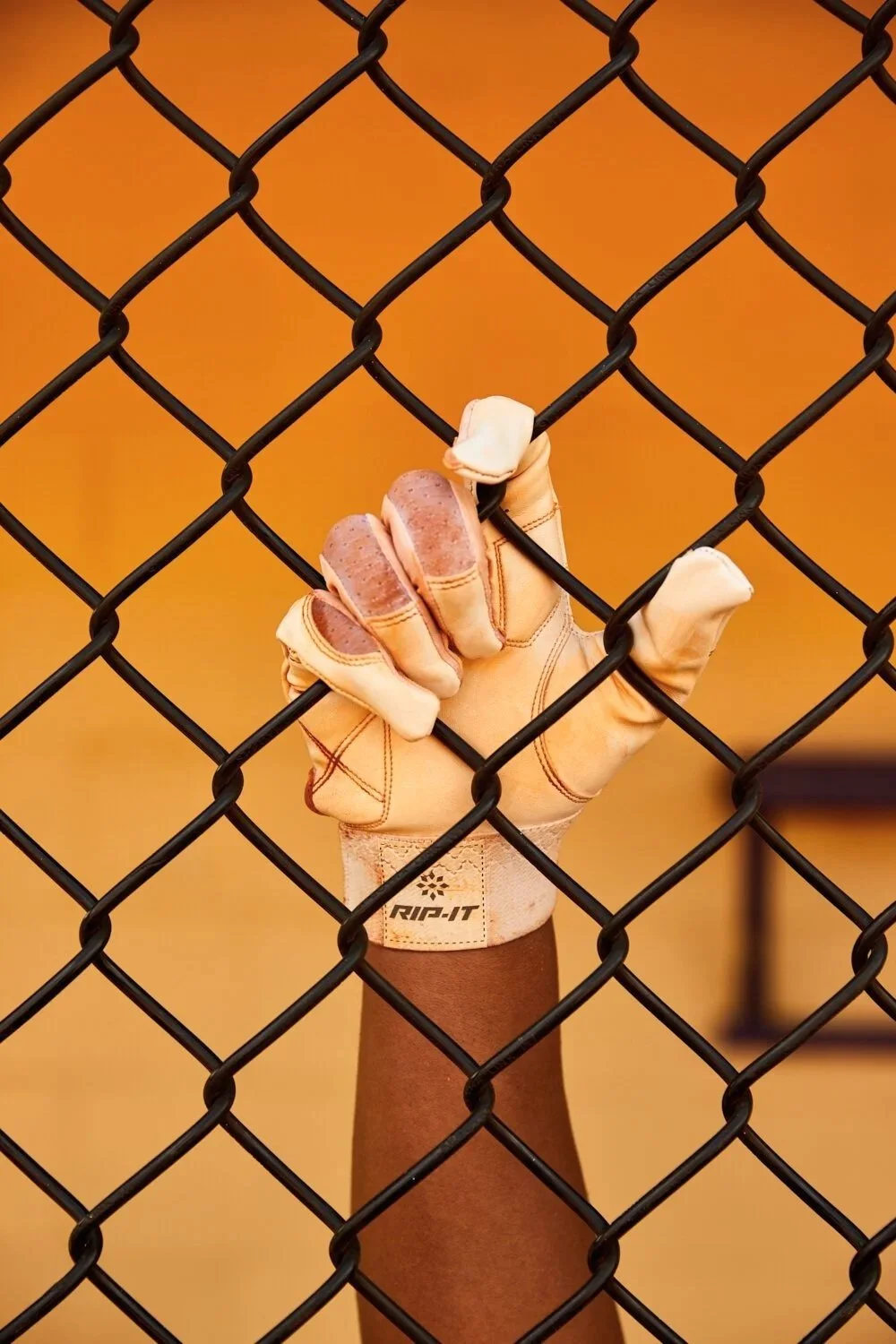 Rip-It Sports - A person wearing a tan leather golf glove gripping a black chain-link fence against an orange background.