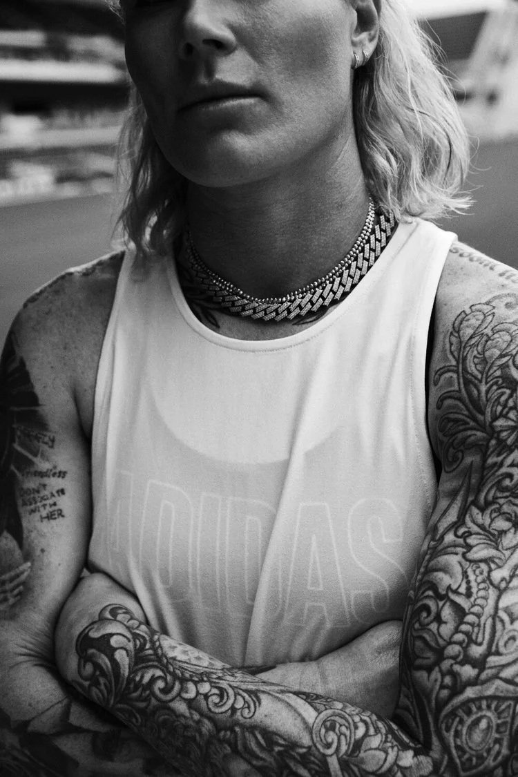 Adidas Photoshoot - A woman with shoulder-length blonde hair and tattoos on her arms wears a sleeveless shirt and a thick chain necklace, standing outdoors in a black and white photo.