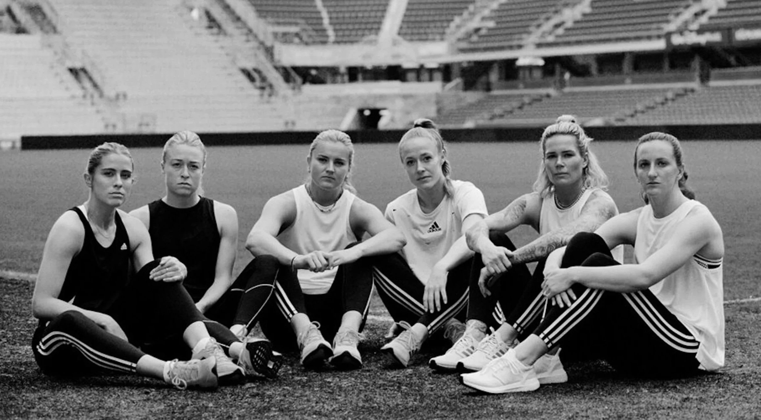 US National Womens Team for Adidas sitting on a track field in a stadium, dressed in athletic wear, posing for a group photo.