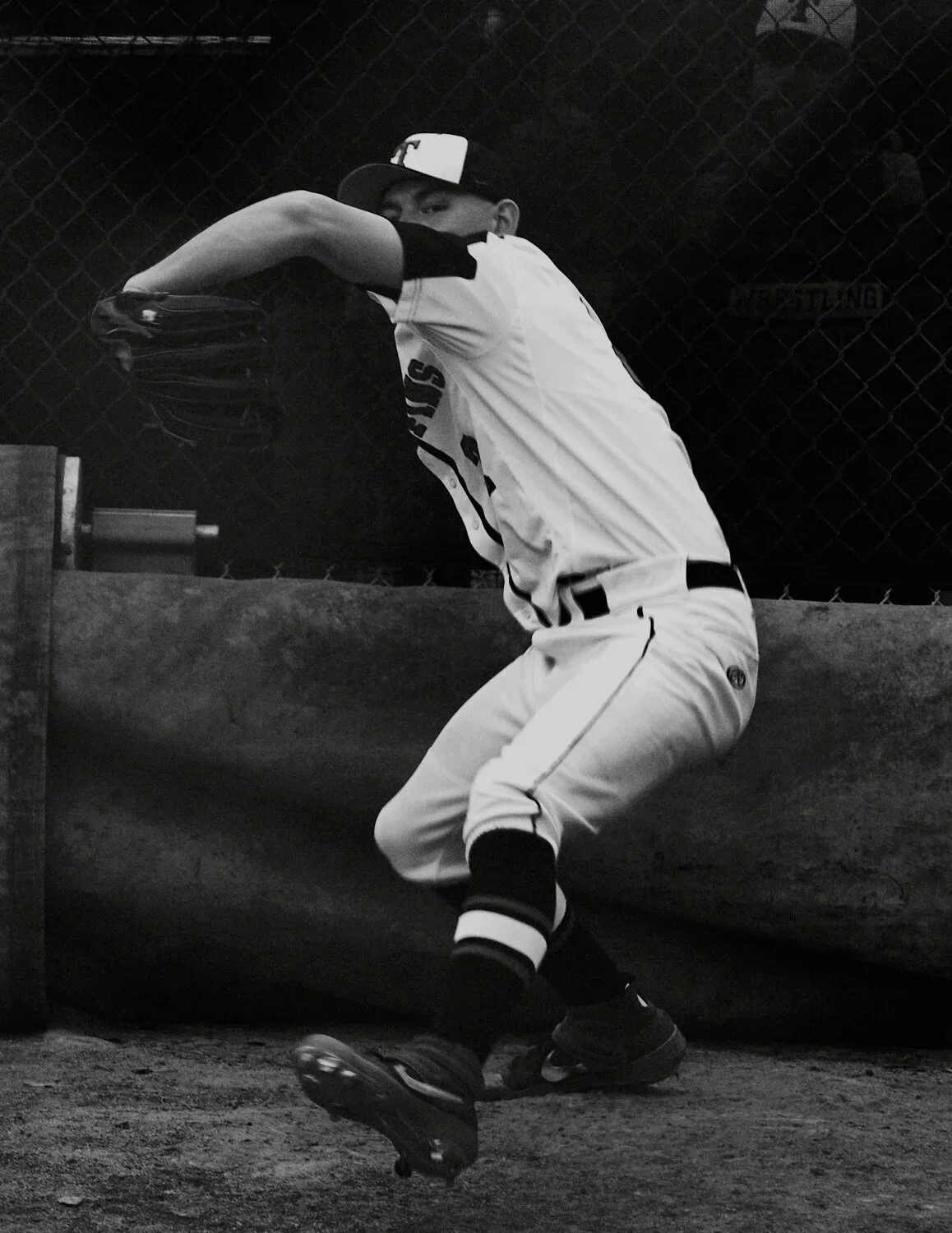 A baseball player in a uniform and cap crouching, wearing a glove, near a fence at night.