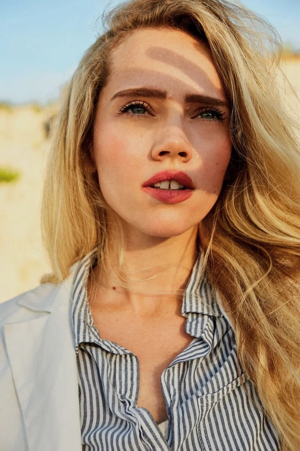 Close-up of a young woman with blonde hair and green eyes, outdoors during golden hour, with sunlight casting shadows on her face.