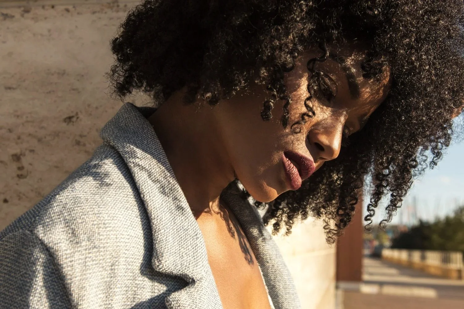 Close-up of a woman with curly hair wearing a grey blazer, looking down against a textured beige wall outdoors during daylight.