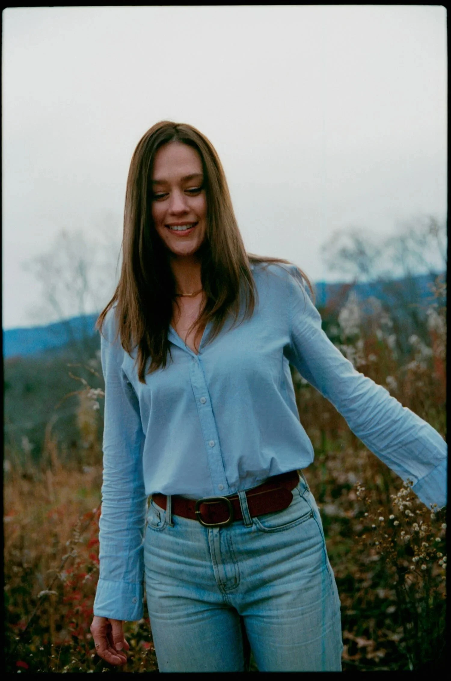 A woman with long brown hair, wearing a light blue button-up shirt and light blue jeans, standing outdoors in a natural setting with trees and mountains in the distance, during overcast weather.
