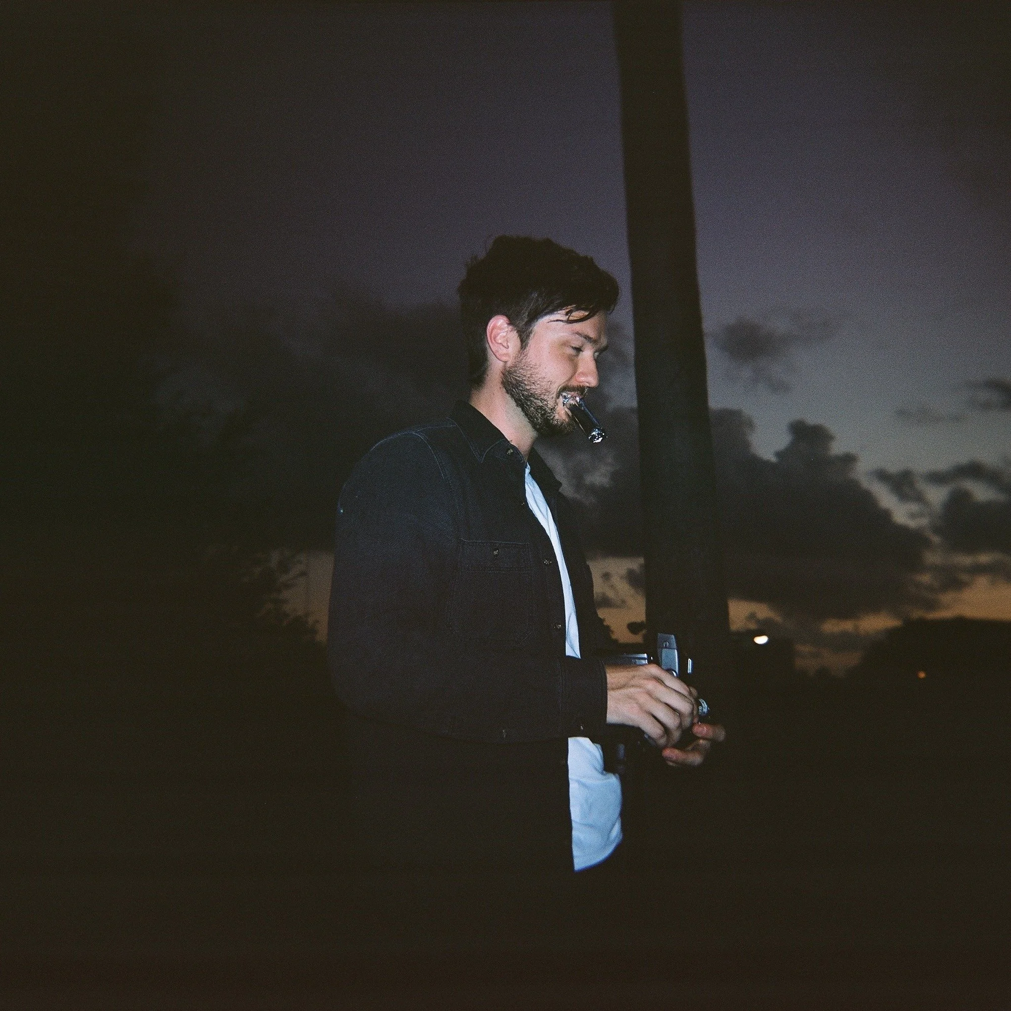 A man with dark hair and a beard wearing a dark jacket and white shirt, holding a camera and a cigarette, standing outdoors during dusk or dawn with silhouetted trees and cloudy sky in the background.