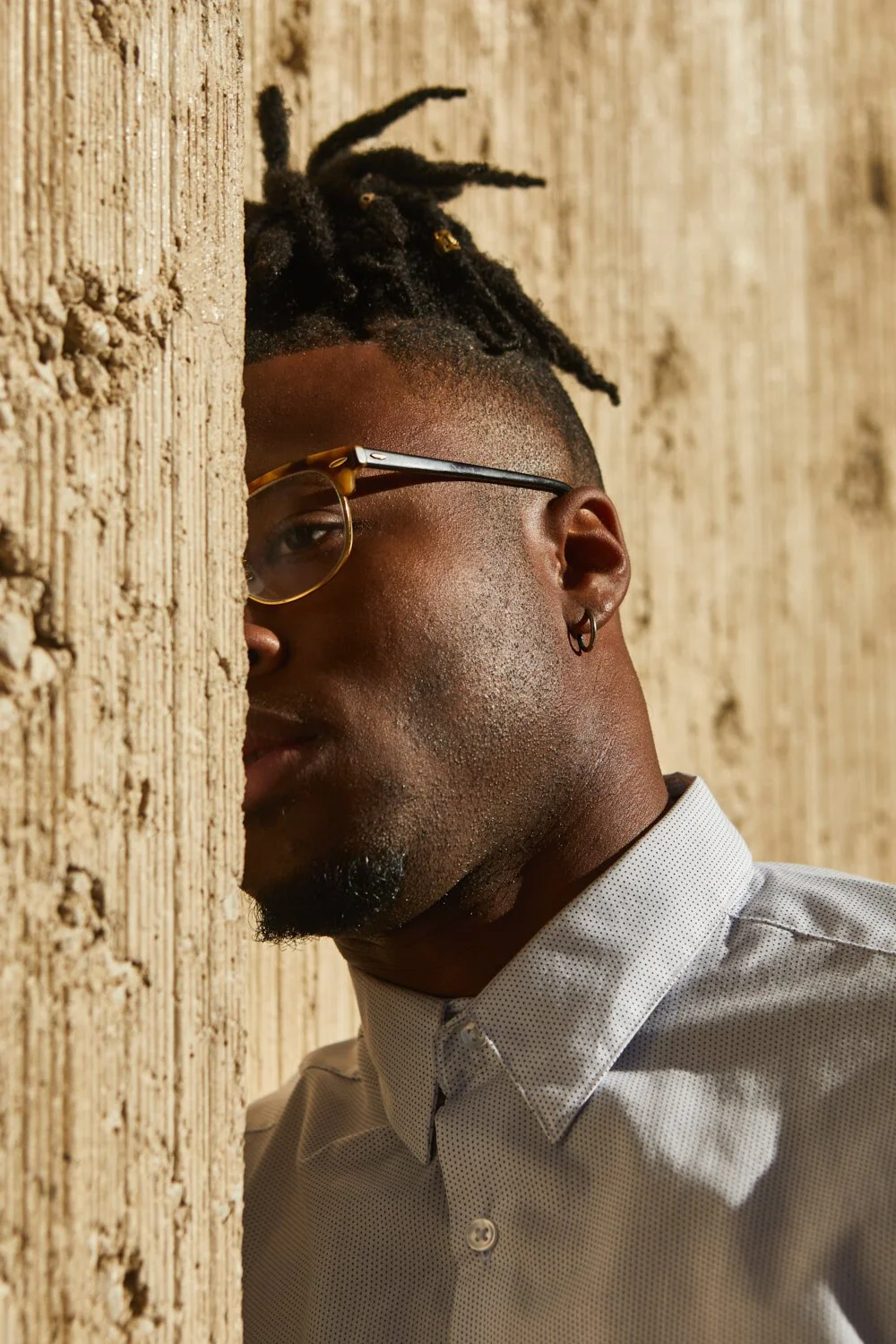 A person with glasses and earrings peeking from behind a textured tan wall, wearing a collared shirt.
