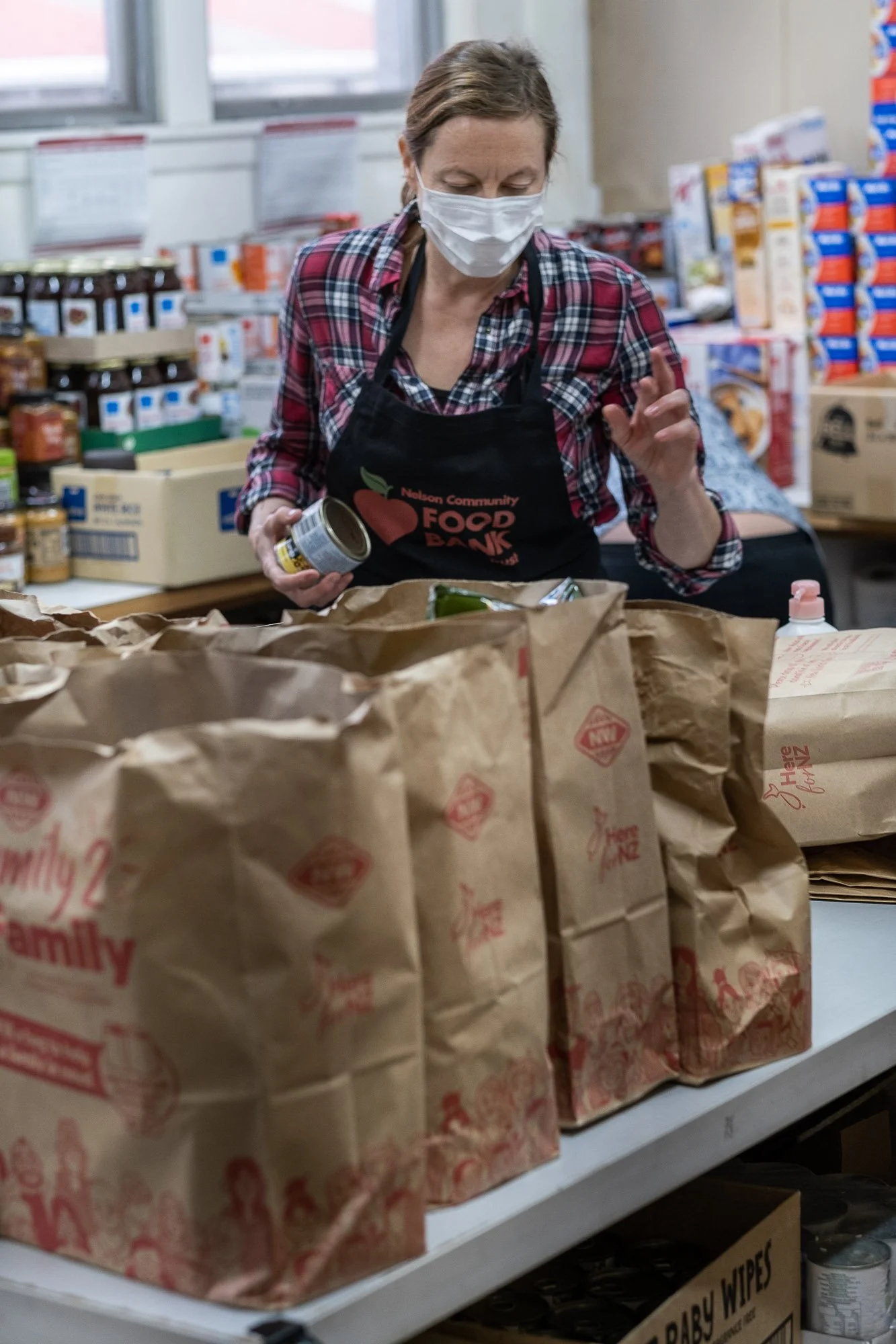 A woman wearing a face mask and an apron working at a food bank, organizing bags of food on a table in a pantry with shelves of canned goods and packages in the background.