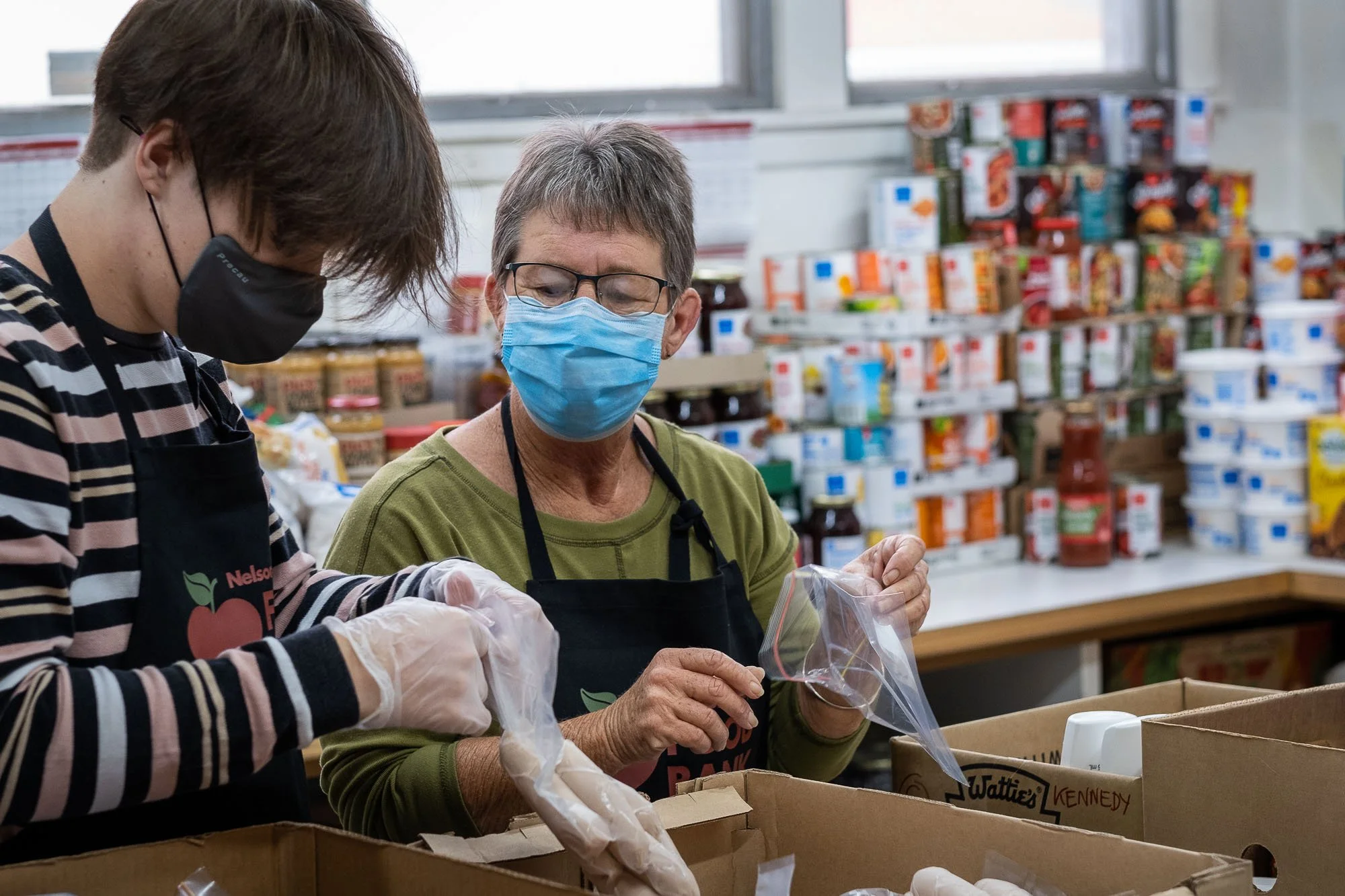 Two women wearing face masks and gloves working together to pack food donations in a food bank, with shelves of canned goods and jars in the background.