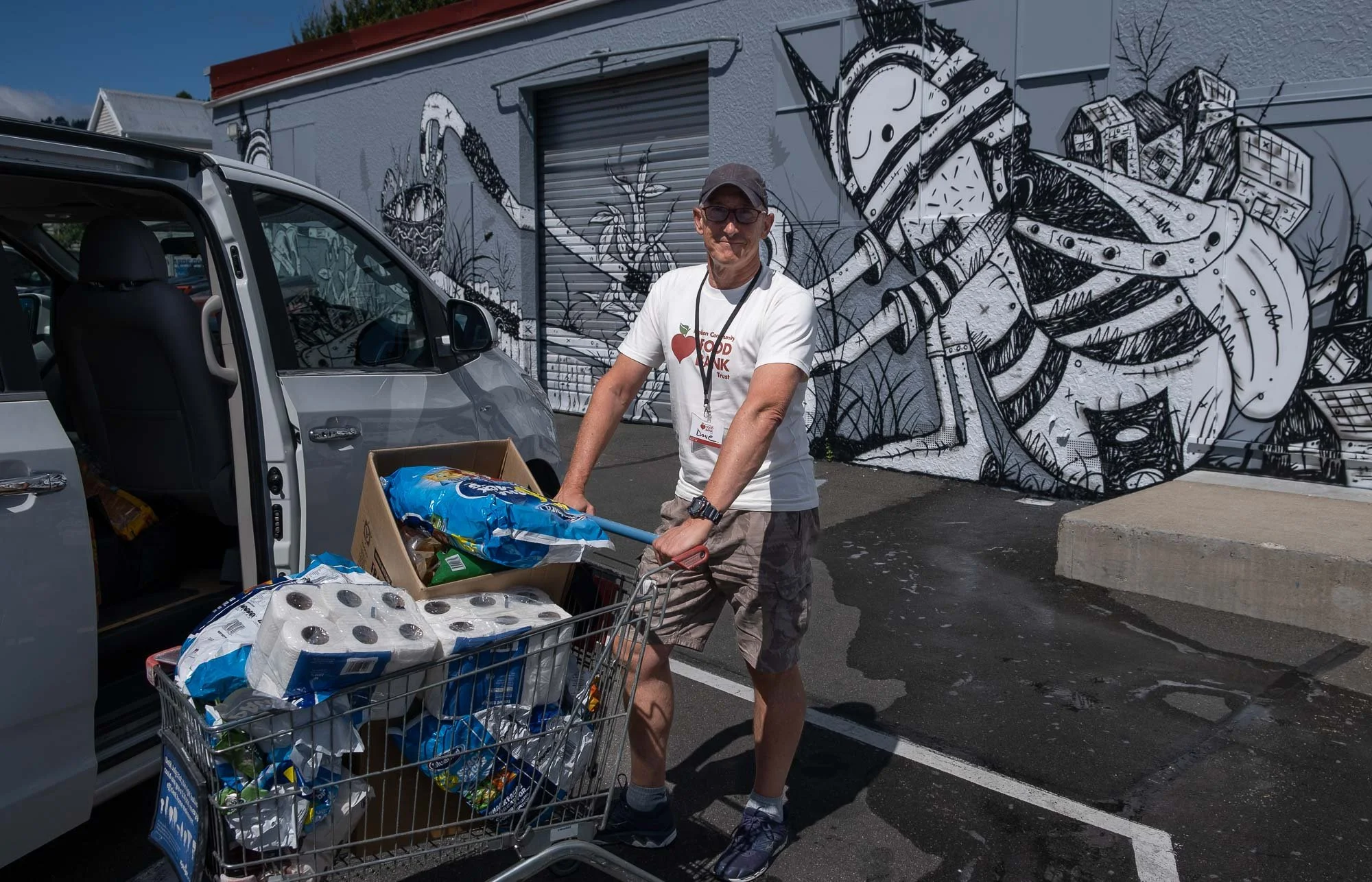 A man in a white t-shirt and shorts filling a shopping cart with items outside a store, with a mural of cartoonish robots and buildings in black and white on the wall behind him.