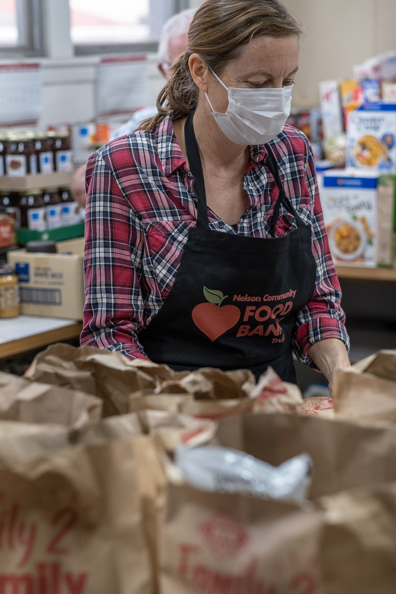 A woman wearing a face mask and a black apron with a heart and leaf logo that reads 'Nelson Community Food Bank' sorts or prepares food at a food bank. She is in a room with shelves of canned goods and cereal boxes.