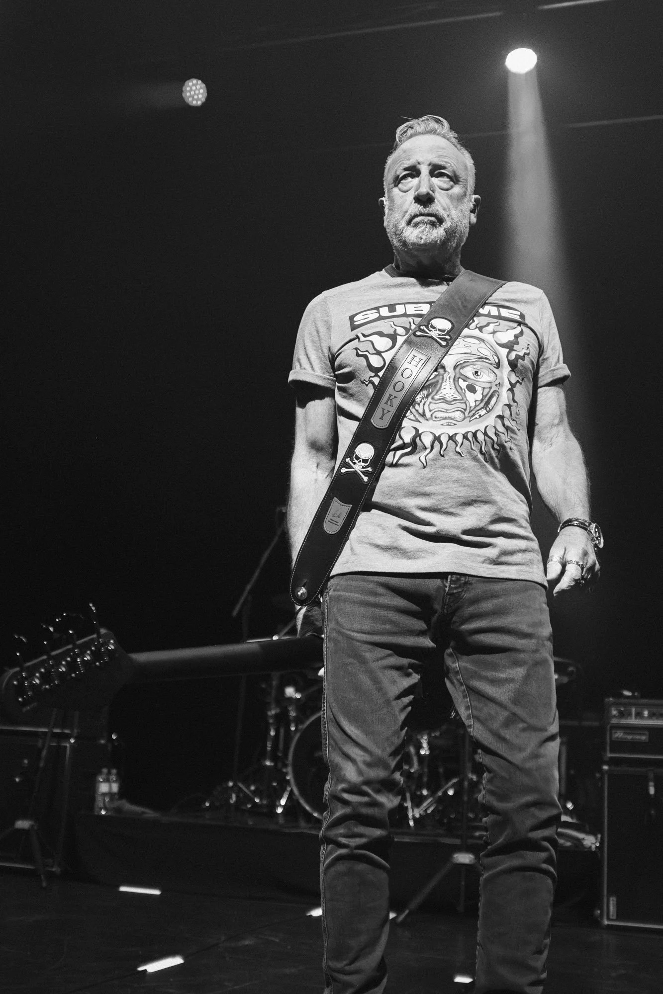 Black and white photo of an older man with a beard standing on stage, illuminated by a spotlight, wearing a graphic t-shirt and jeans, with musical equipment in the background.