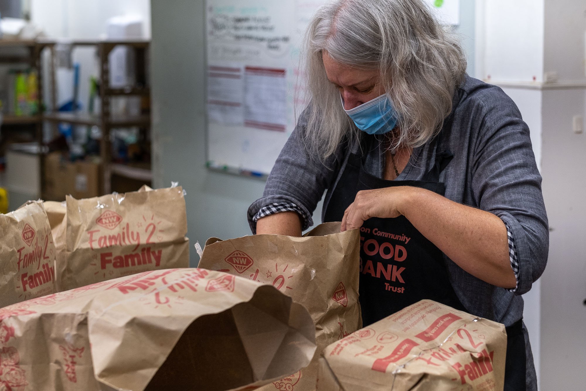 Woman wearing a face mask and black apron packing food into paper bags at a community food bank.