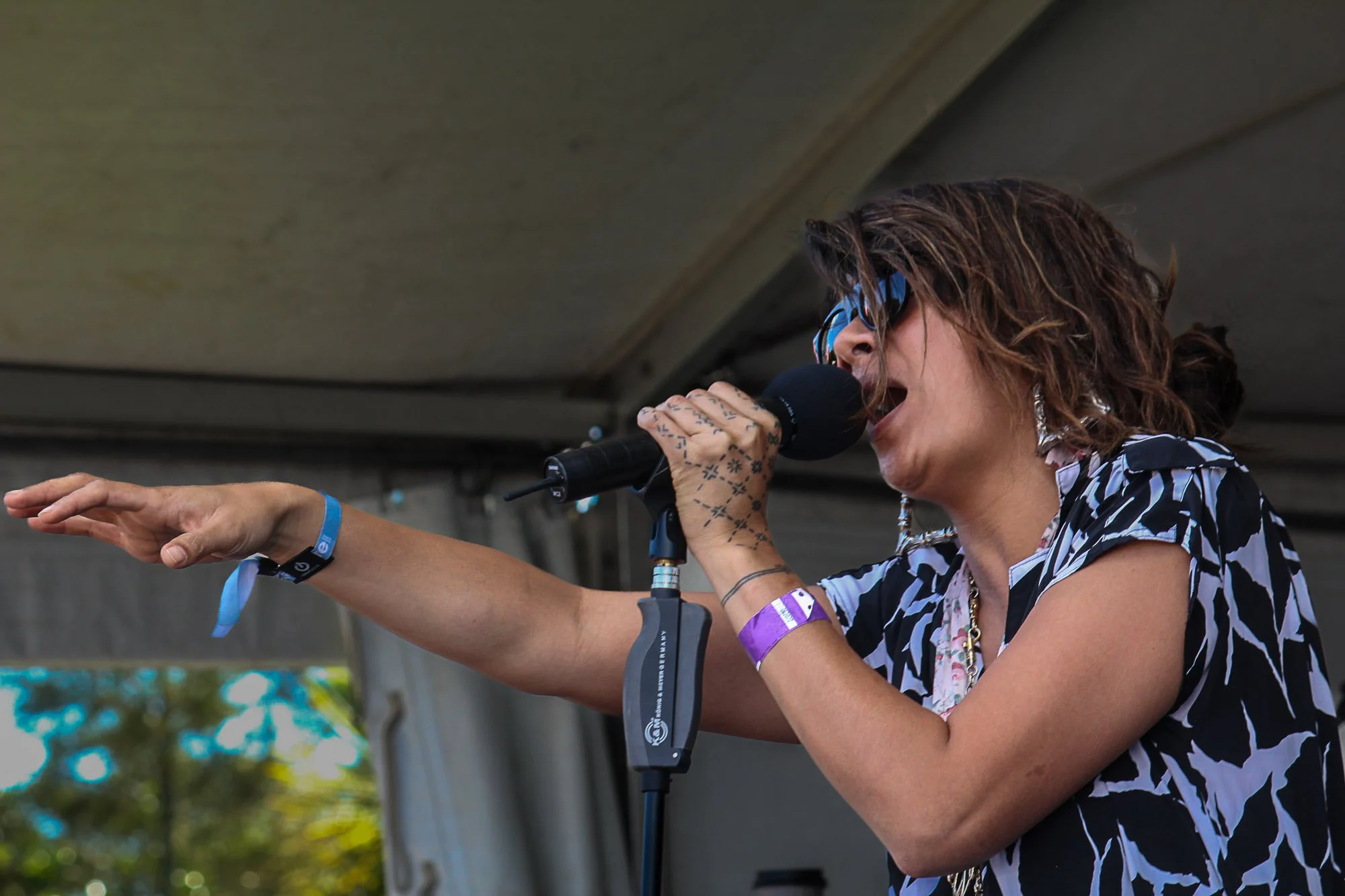 A woman singing into a microphone at an outdoor event, wearing sunglasses, a patterned shirt, and wristbands, with her right arm extended.