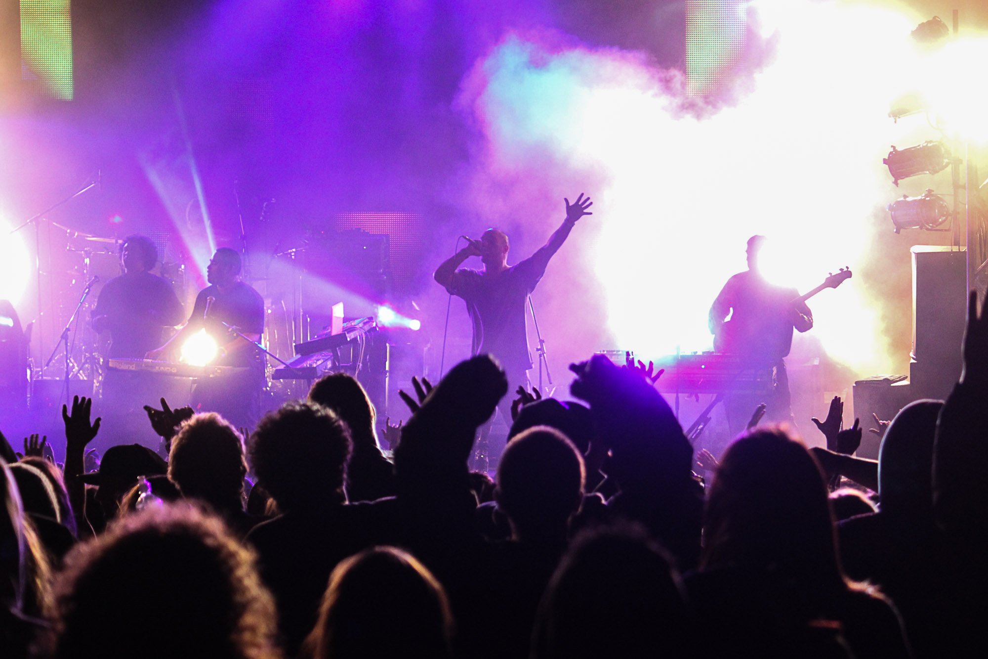 A live concert scene with band members on stage amid colorful purple, yellow, and blue lighting and fog effects. The audience is in front with hands raised.
