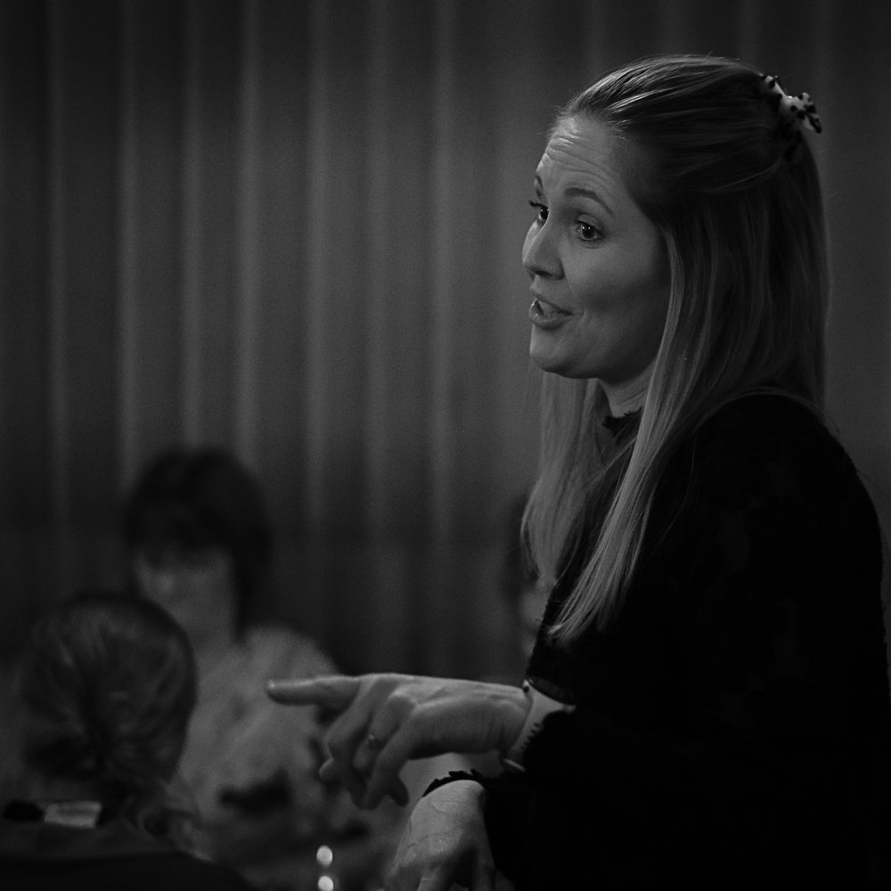 A woman with long hair, speaking and gesturing, with a blurred background of two children sitting and listening.