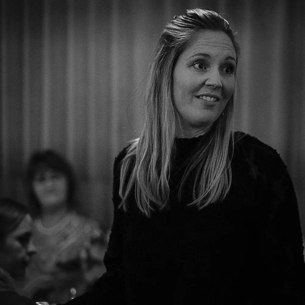 A woman with long hair smiling, wearing a dark top, with two other women blurred in the background, in a room with curtains.