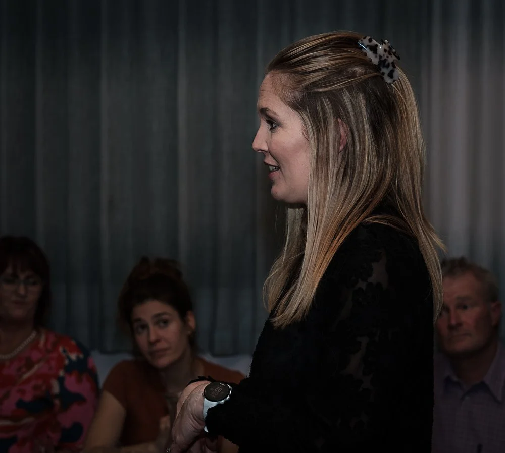 A woman with long blonde hair, wearing a black outfit and a leopard-print hair clip, speaking to a group in a dimly lit room with dark curtains. Seated audience members are visible in the background.