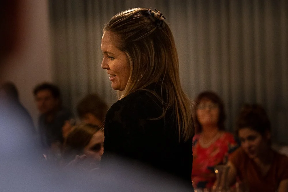 A woman with long brown hair, smiling, talking to others at a social event, with a blurred group of people in the background.