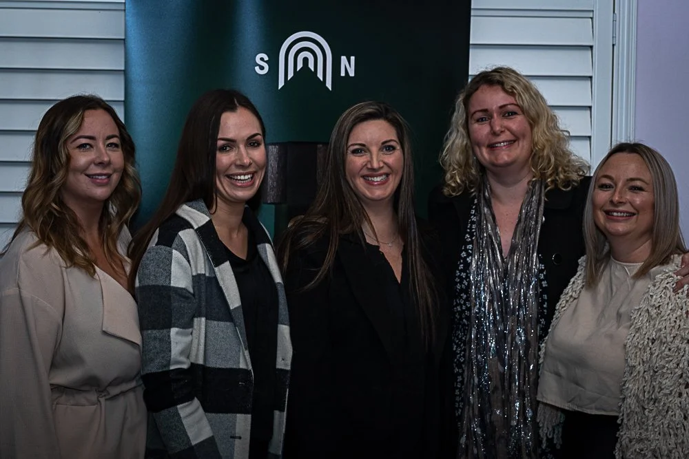Five women standing together, smiling for a photo, in front of a banner with a rainbow and the letters 'S' and 'N' on it.