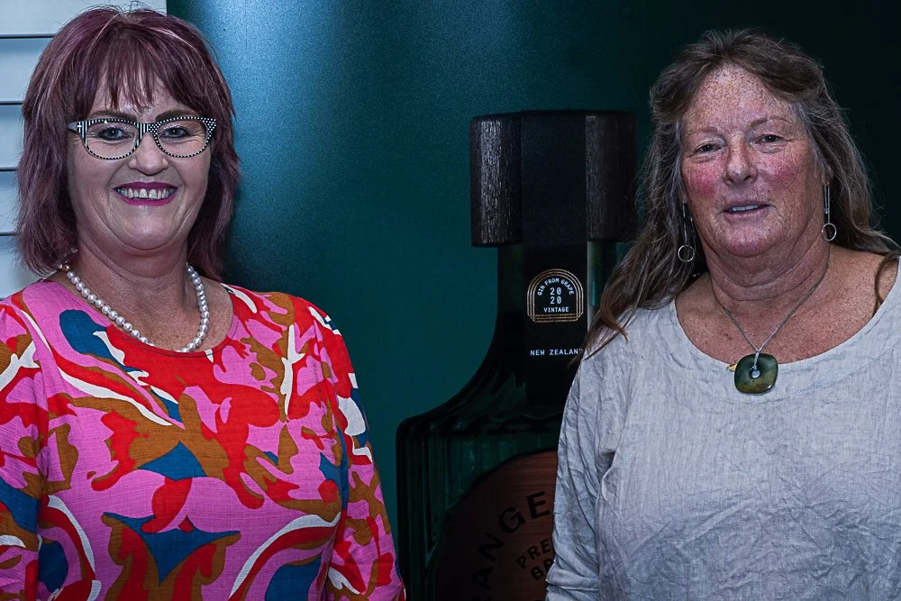 Two women standing indoors, smiling at the camera, with a lamp and a bottle of something behind them.