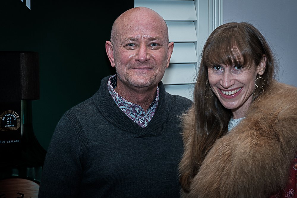 A smiling man with a shaved head and a woman with long brown hair and bangs smiling, standing indoors close to each other.