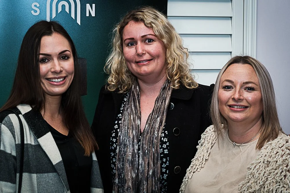 Three women standing together indoors, smiling at the camera.