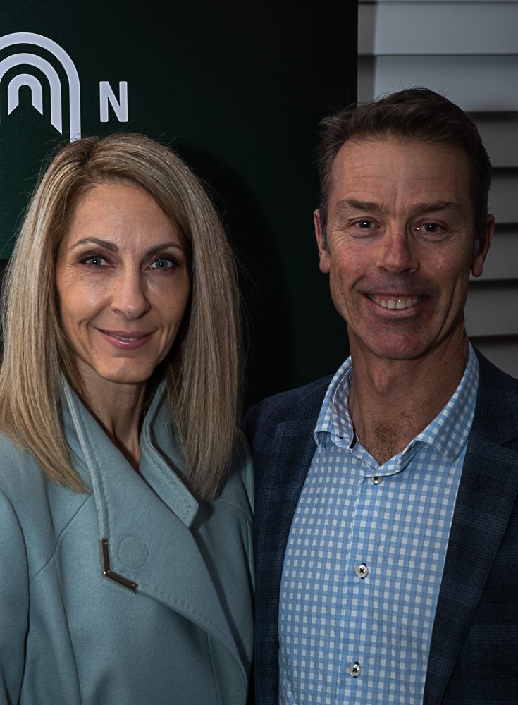 A woman and a man standing close together, smiling for the camera. The woman has blonde hair and is wearing a light blue coat, and the man has short brown hair and is wearing a blue checkered shirt with a dark blazer. They are indoors, with a dark ba