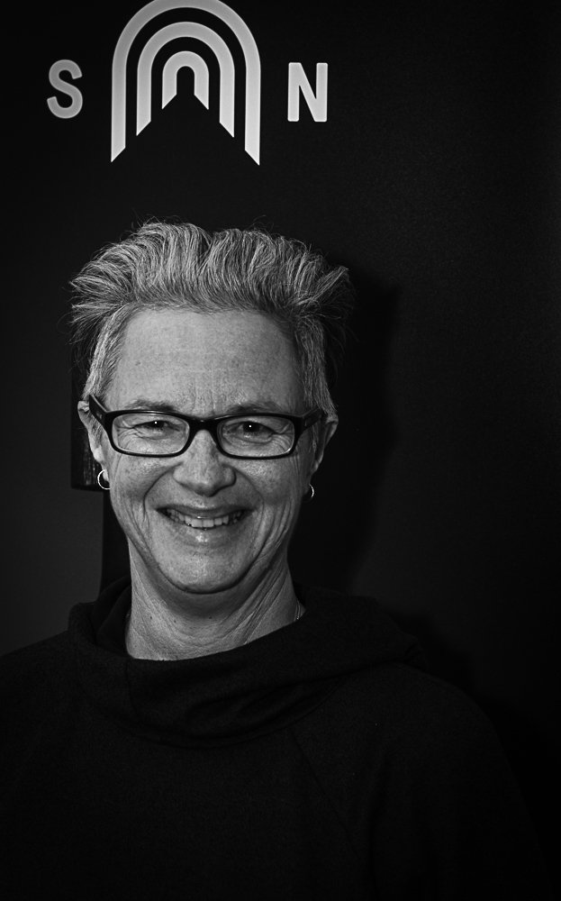 A woman with short, styled hair and glasses smiling in front of a black background with a white logo and the letters S and N.