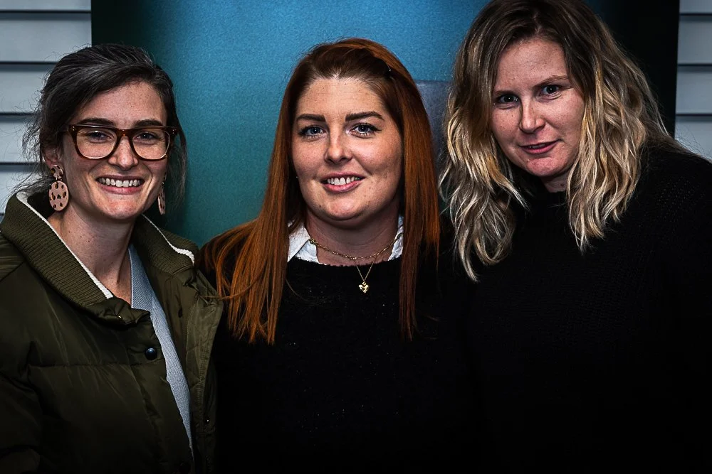 Three women standing close together, smiling, against a blue wall with window blinds in the background.