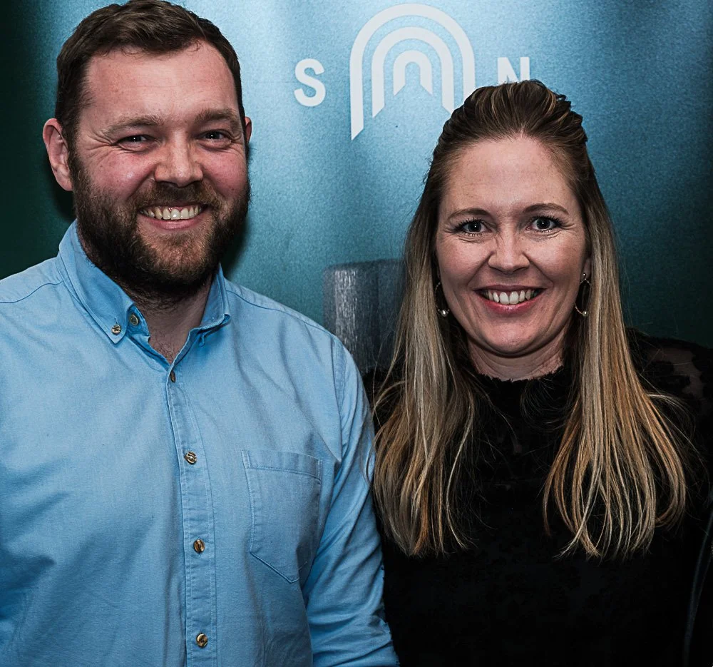 Two smiling people standing in front of a blue background with a logo and the letters 'S' and 'N' visible.