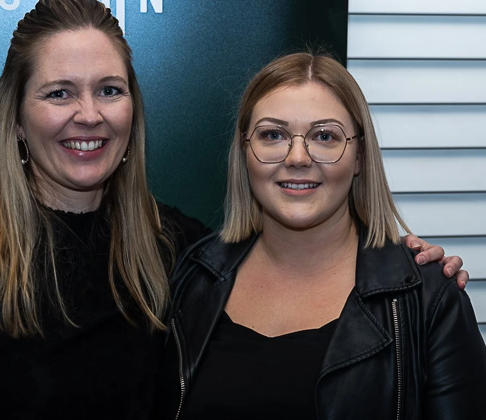 Two women smiling and standing close together, with one woman having her hand on the other's shoulder. They are indoors, with a blue wall and white louvered panel in the background.