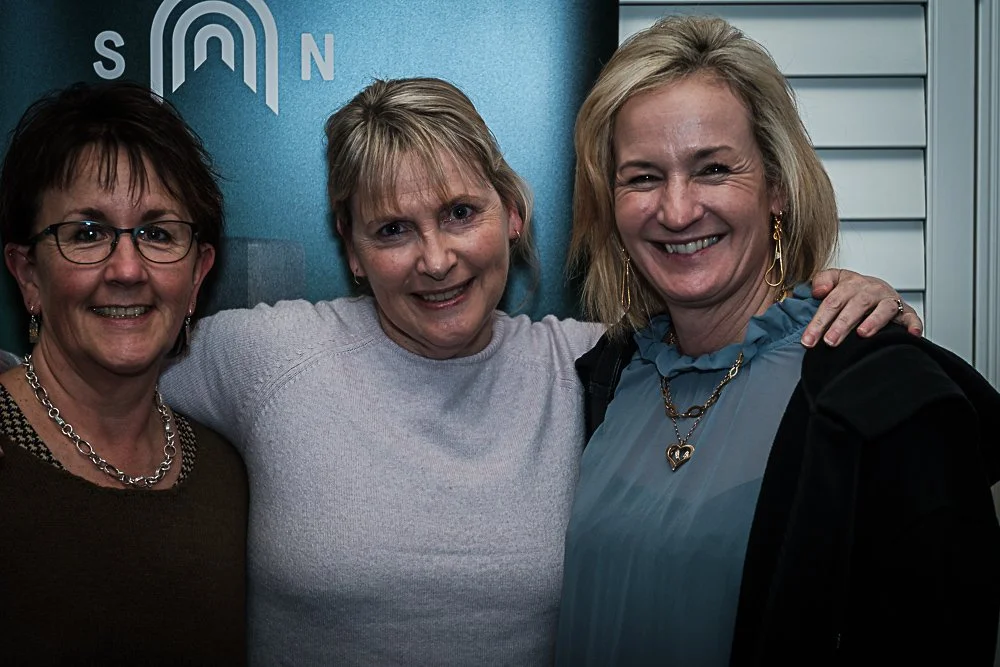 Three women smiling and standing close together indoors, with one woman resting her arm on another's shoulder.