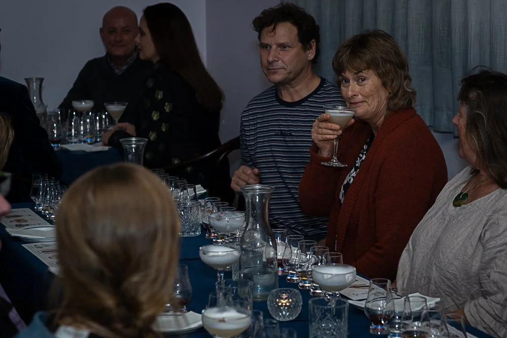 A group of people gathered around a table at a social event, with several glasses, bottles, and trays of drinks.