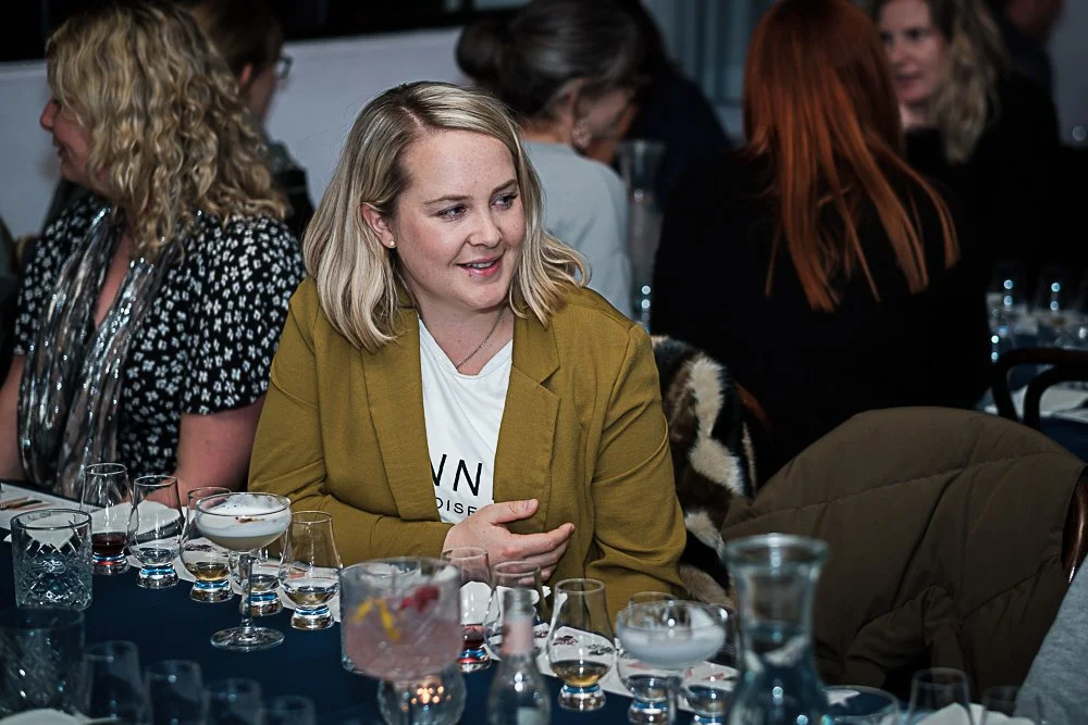 A woman with blonde hair wearing a yellow blazer is sitting at a table with various glasses of drinks, in a crowded restaurant or event setting.