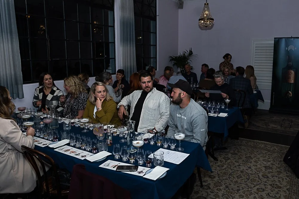 People participating in a whiskey or liquor tasting event at a banquet table in a dimly lit room with large windows, curtains, and a chandelier.