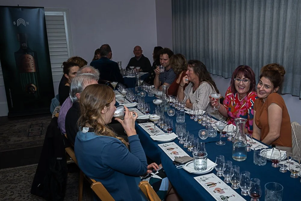 People sitting at a long table with glasses of drinks and tasting items, engaged in conversation at a social gathering or tasting event.