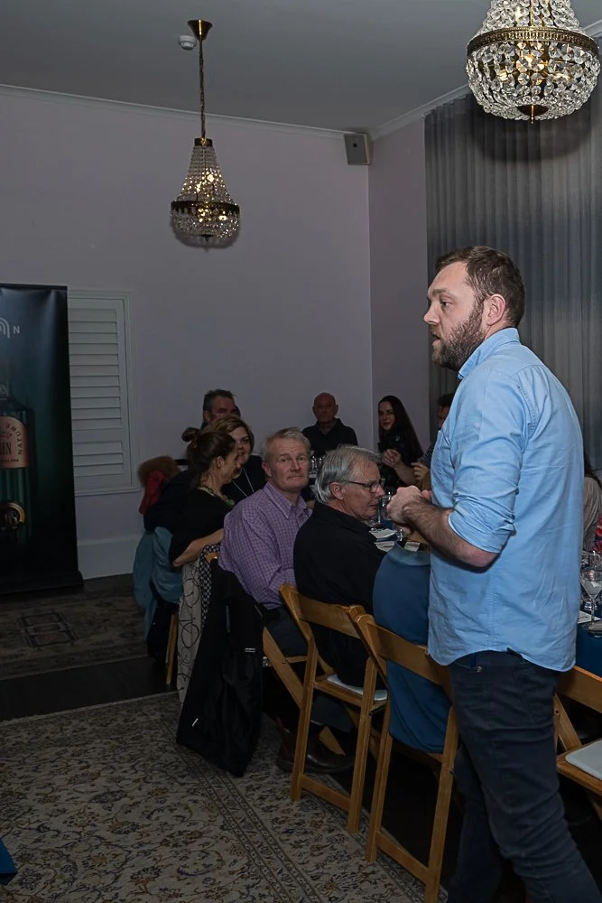 A man with a beard and wearing a light blue shirt stands at the head of a dinner table, speaking to a group of seated guests in a room with chandeliers and gray curtains.