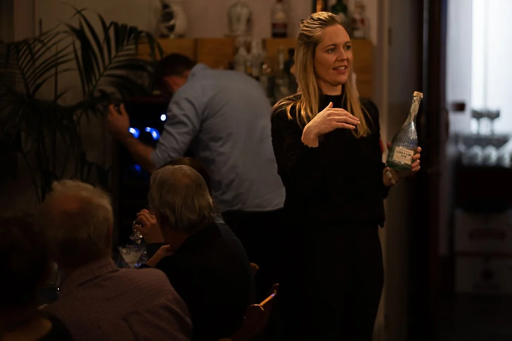 A woman holding a bottle of water or clear beverage, standing and talking in a dimly lit room with several people seated at tables, and a man in the background working on a computer.