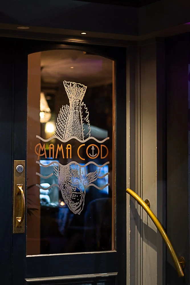 Glass door with a fish graphic and the text 'Mama Cod,' featuring an anchor symbol, with a gold handle and a dark-colored exterior wall.