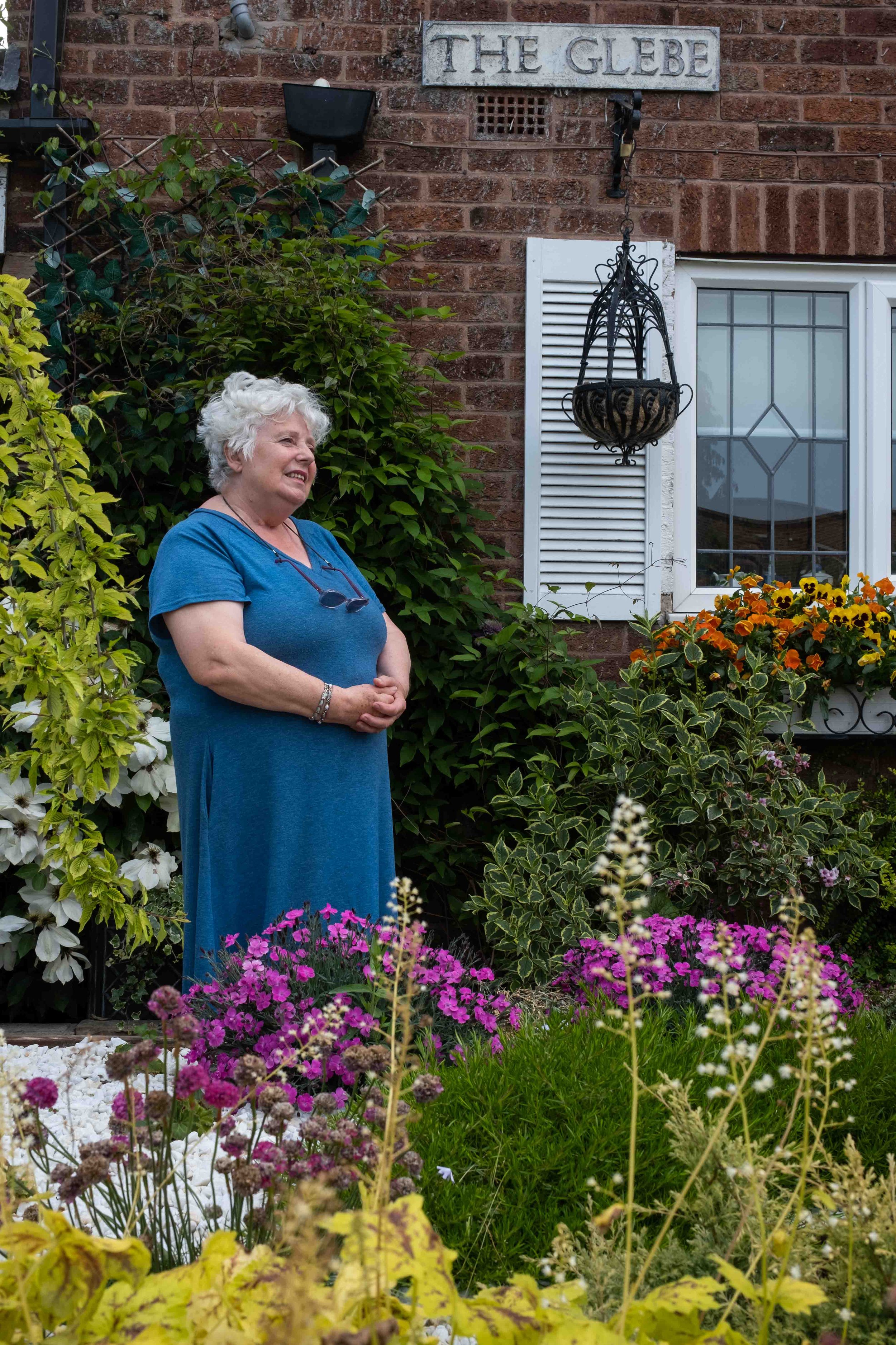 Older woman with white hair wearing a blue dress, smiling and standing in a colorful garden with various blooming flowers in front of a brick house with a sign that reads 'The Glebe'.