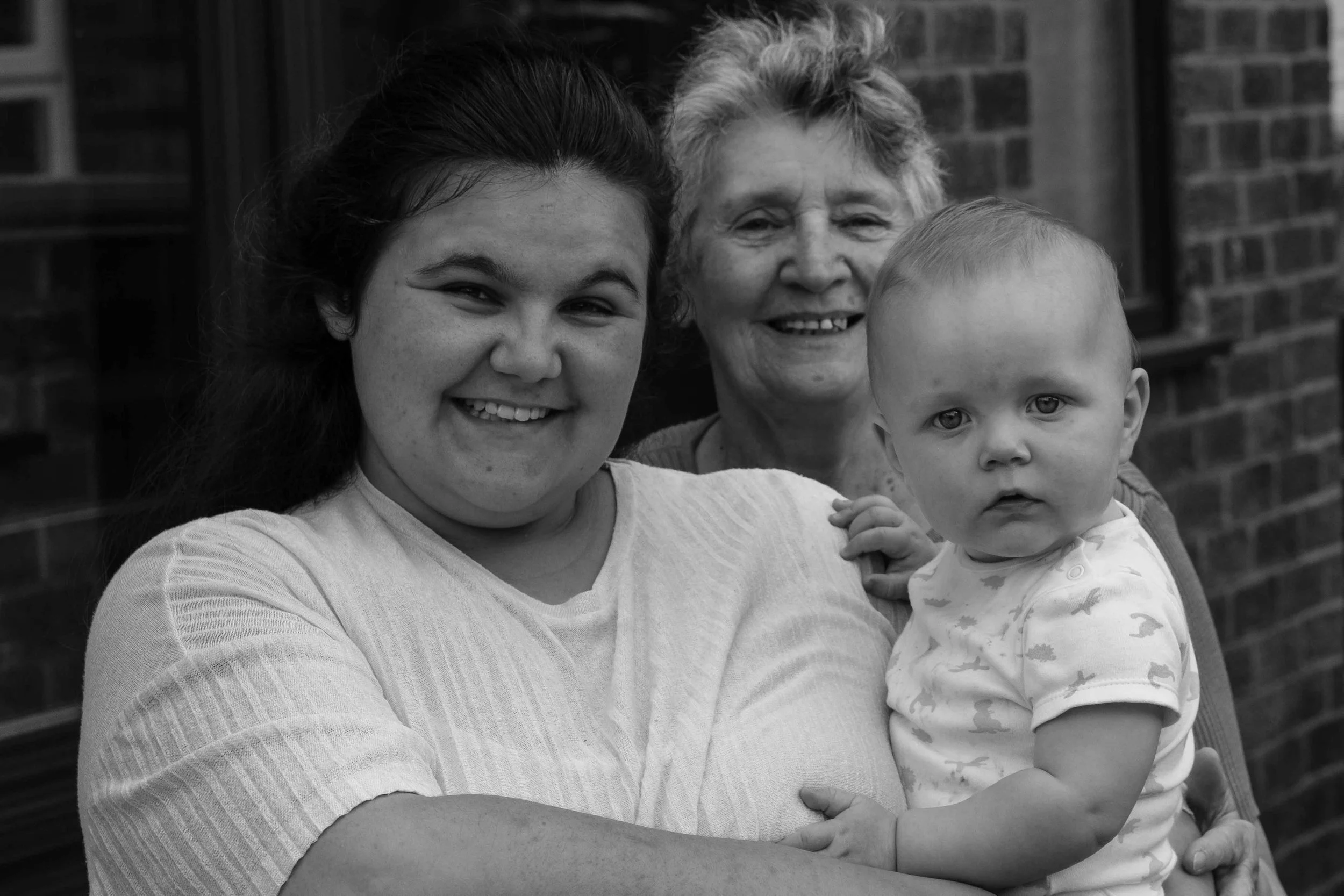 Three generations of women smiling outside, with a brick wall in the background. The woman on the left has dark hair and is holding a young child with light hair, who looks curious. An older woman with short, curly hair is in the center, smiling.