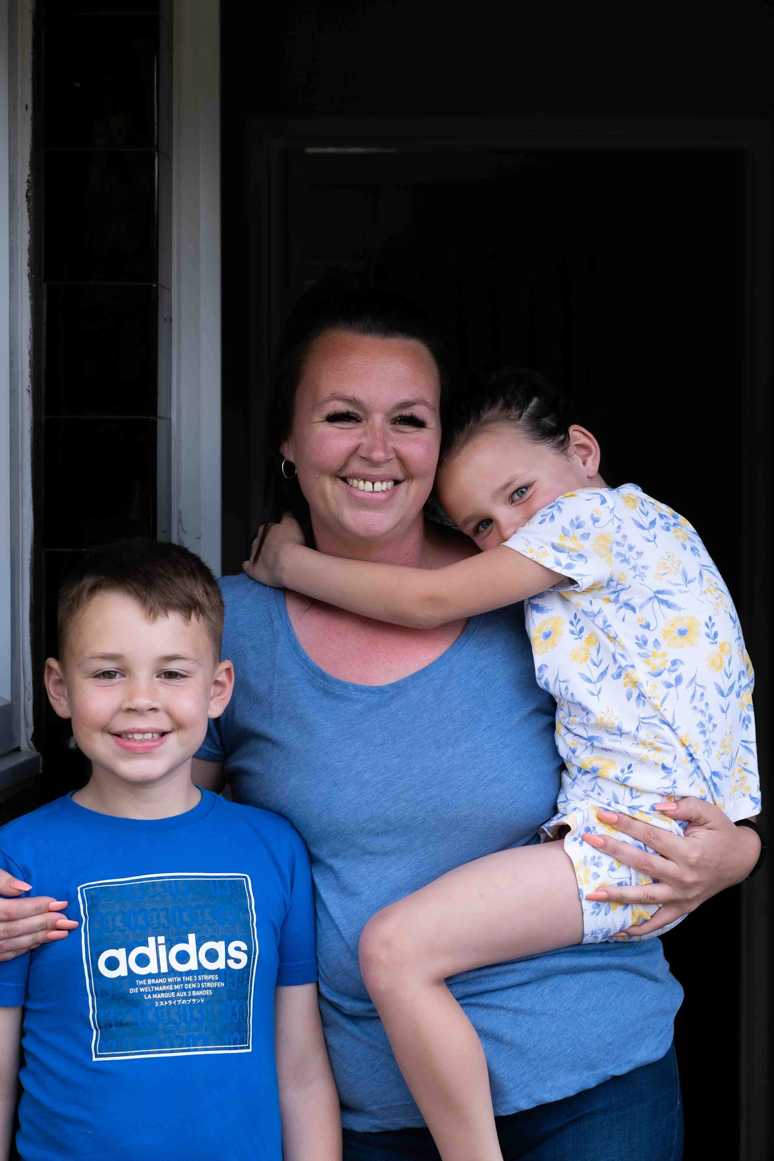 A woman smiling while holding a young girl in her arms and a young boy standing next to her, outside a house.