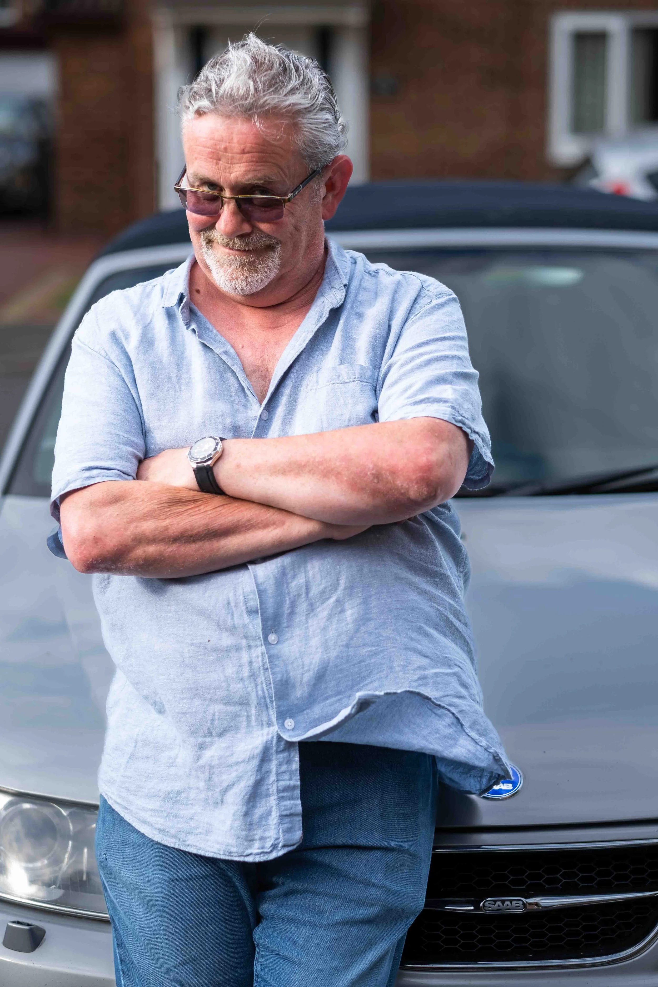 An older man with gray hair, wearing glasses, a light blue short-sleeved shirt, and jeans, stands in front of a silver car with crossed arms, smiling.
