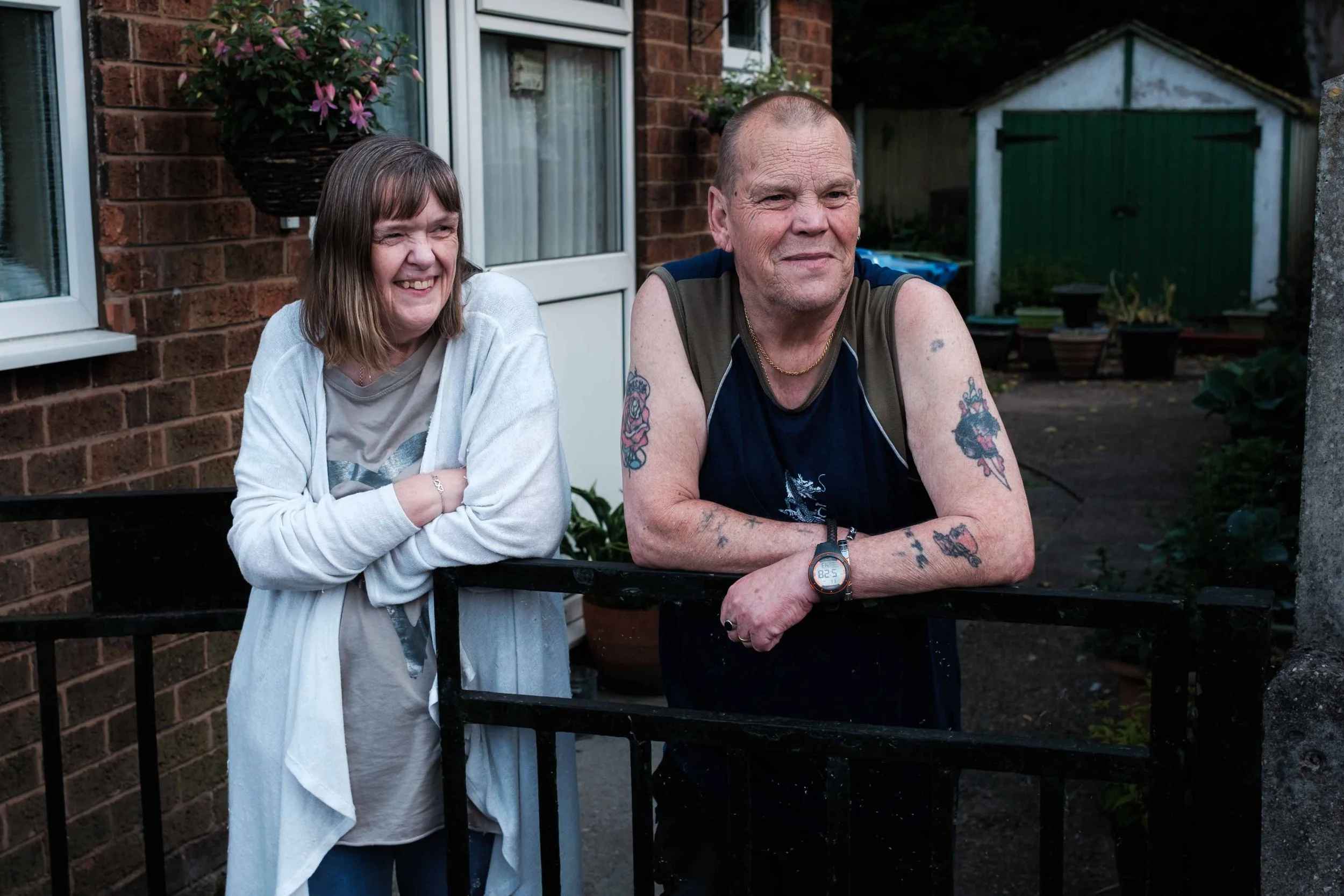 A woman and a man stand outside near a black metal gate, smiling. The woman is wearing a light gray jacket and a T-shirt, while the man has tattoos on his arms and is wearing a sleeveless shirt and a watch. They are in a backyard with a brick house, potted plants, and garden shed in the background.