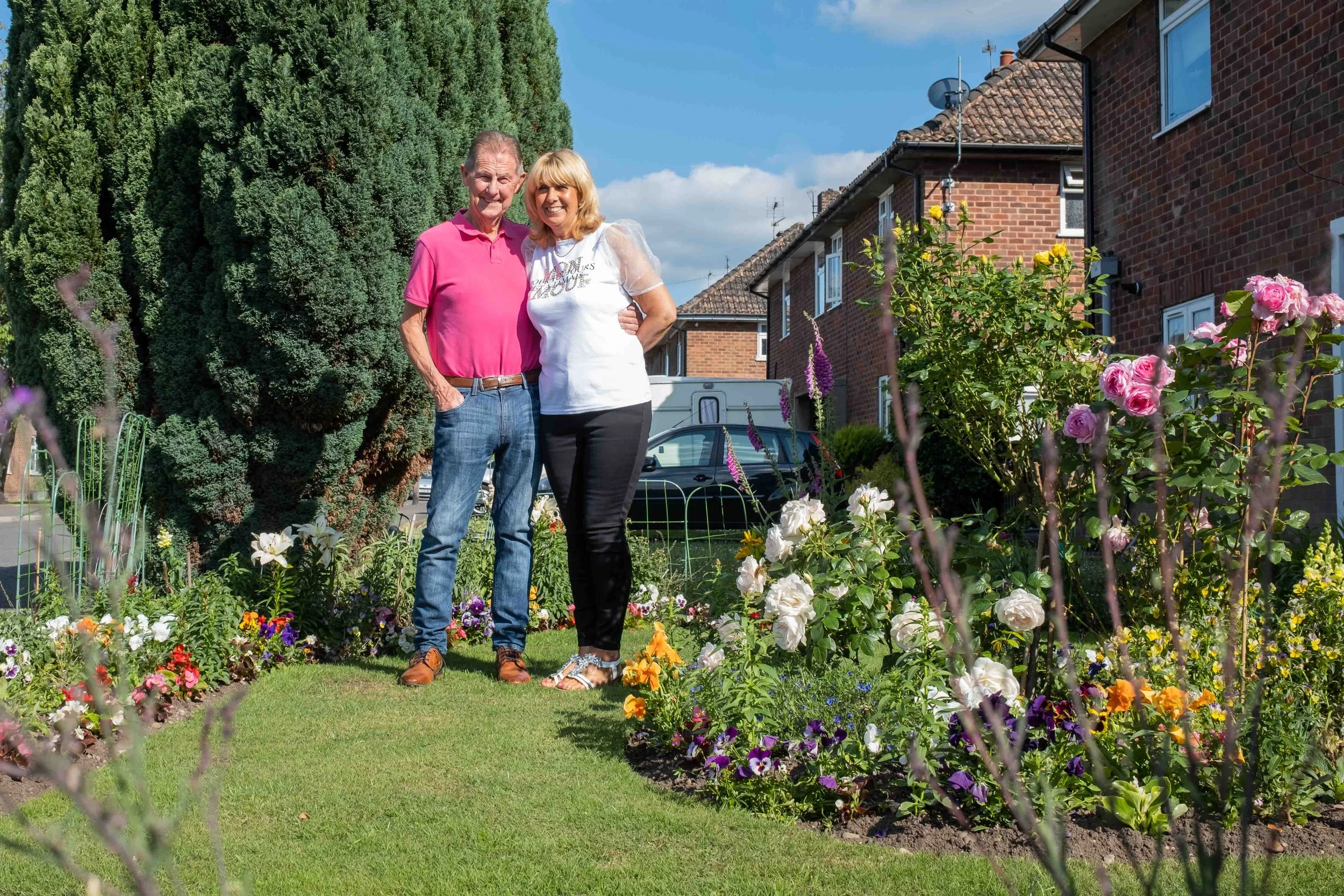 A couple standing together in a garden with colorful flowers, with houses and trees in the background on a sunny day.
