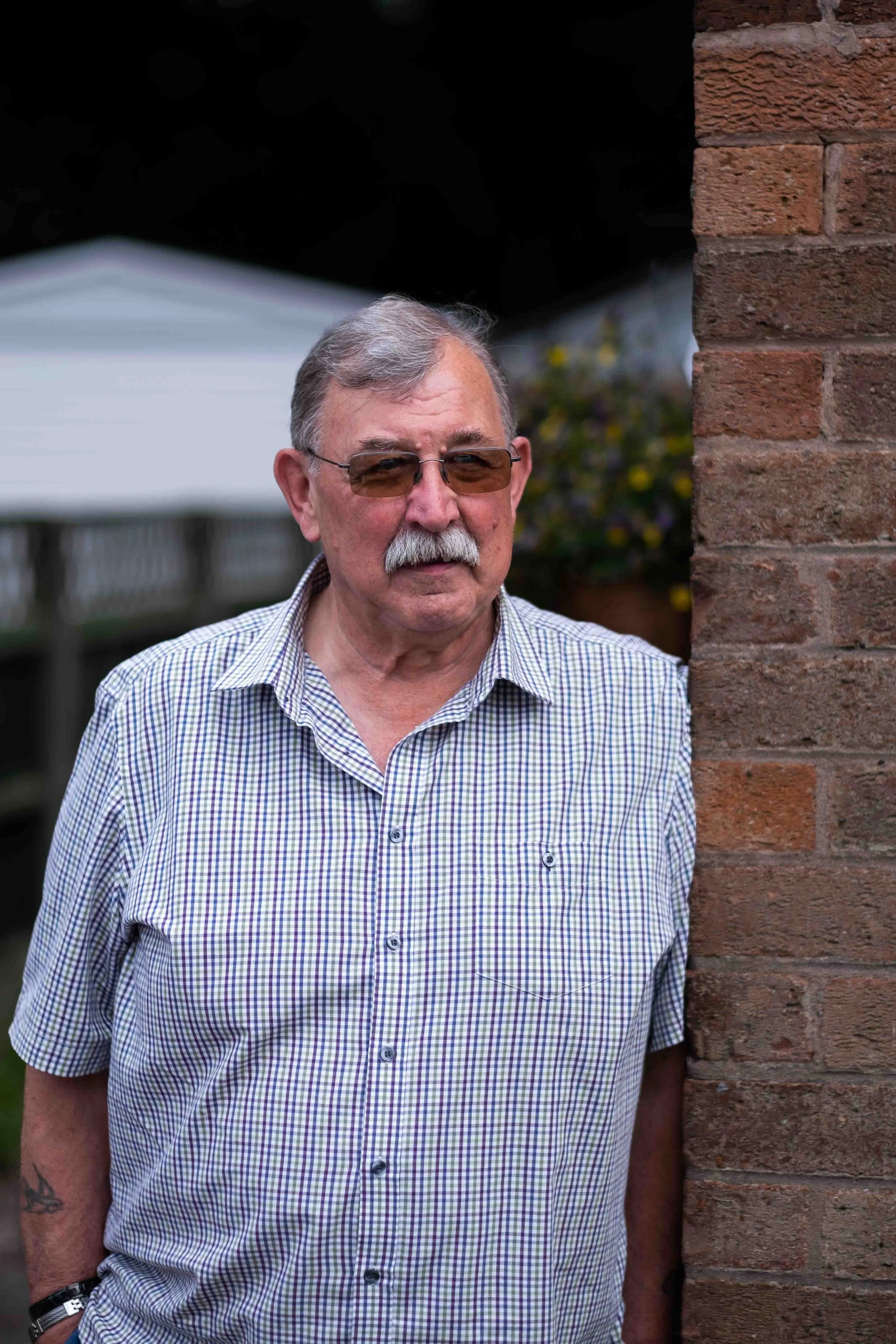 An elderly man with gray hair, mustache, and sunglasses standing beside a brick wall, wearing a short-sleeved, checkered shirt.