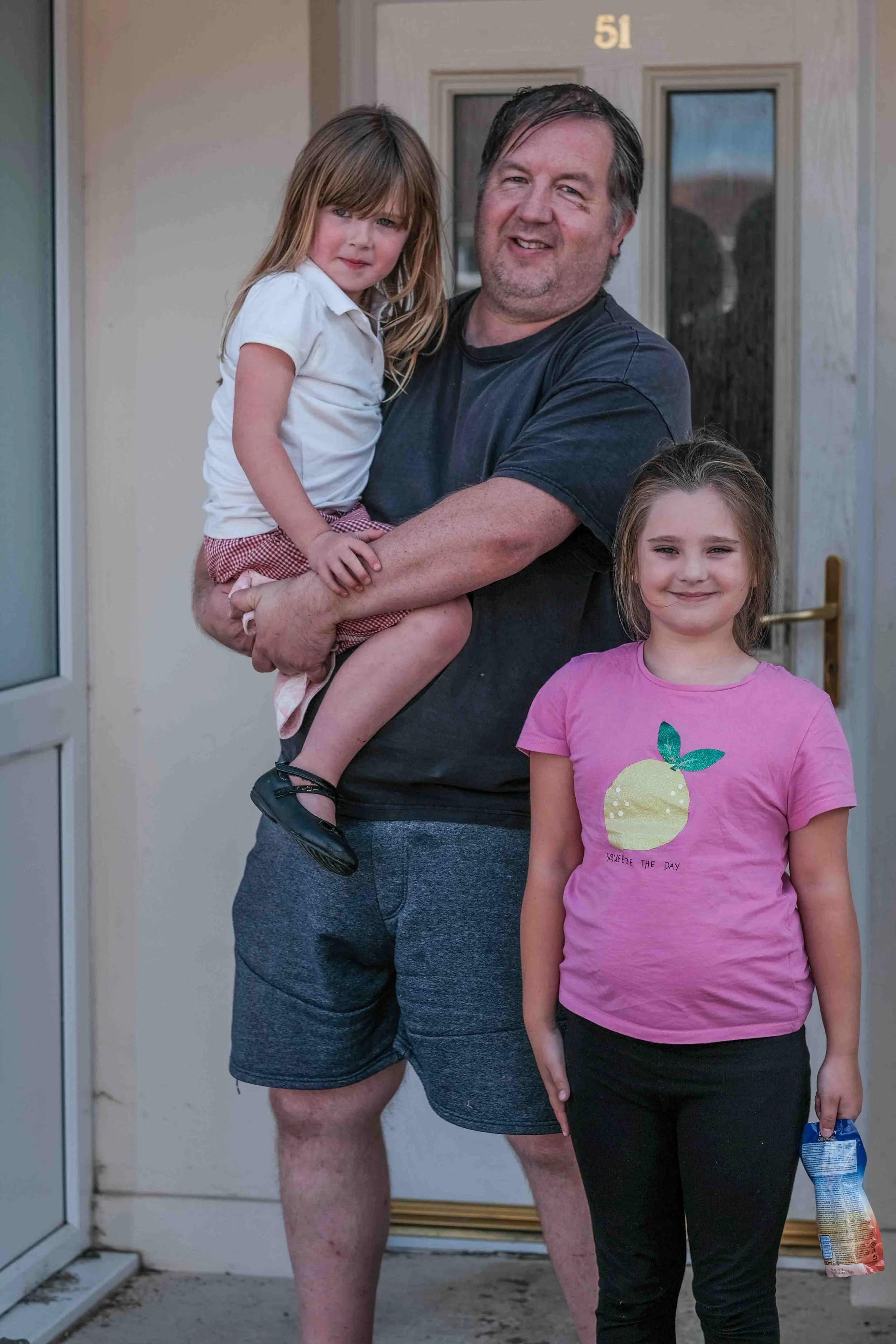 A man holding a young girl in front of a house, with another girl standing beside him. The man is smiling, wearing a black t-shirt and gray shorts. The girl he is holding has long blonde hair, wearing a white shirt and checkered shorts. The girl standing has long hair, wearing a pink t-shirt with a yellow fruit graphic, and black pants, holding a snack bag.