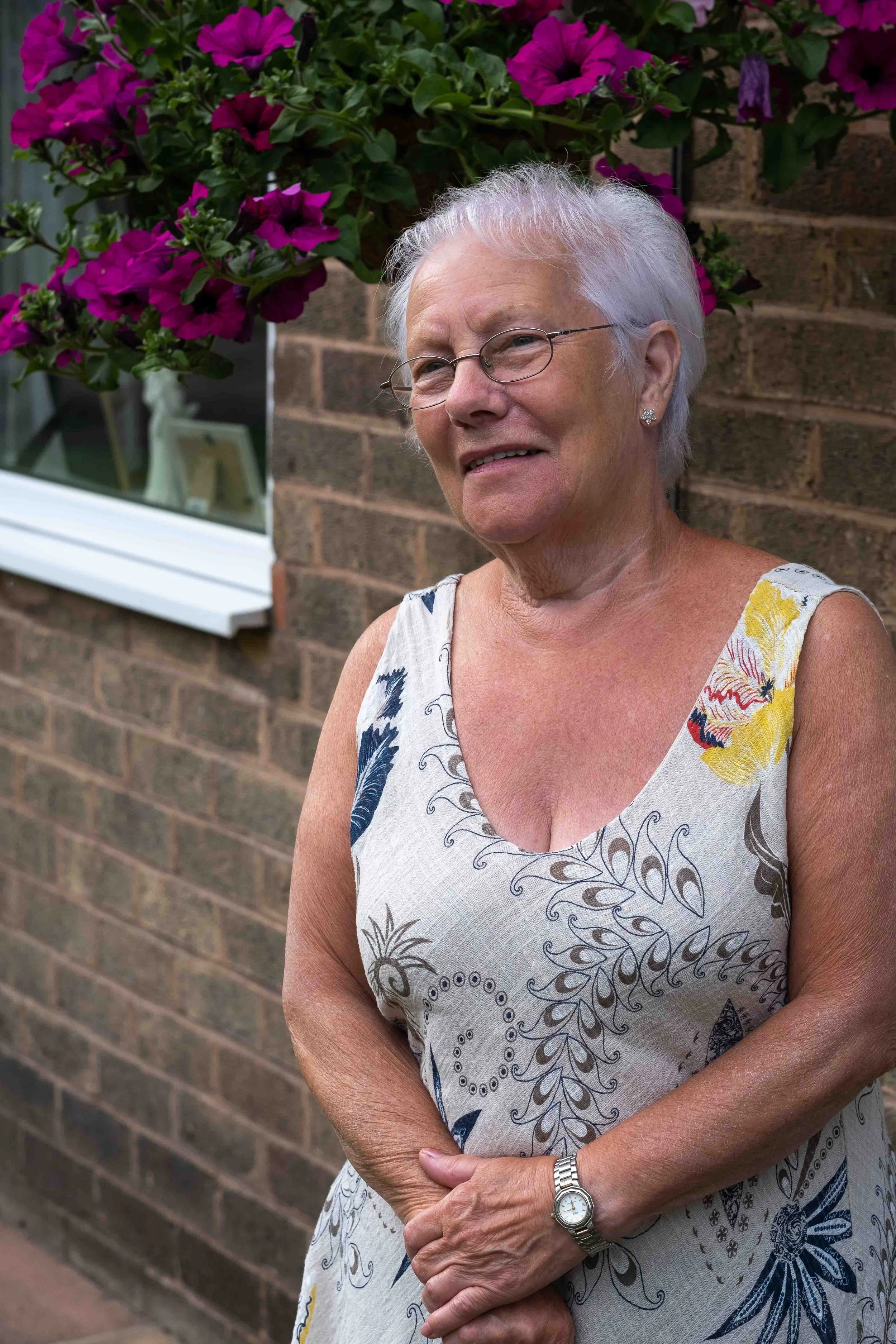 Older woman with white hair and glasses leaning against a brick wall near pink flowers, wearing a sleeveless floral dress and a wristwatch.