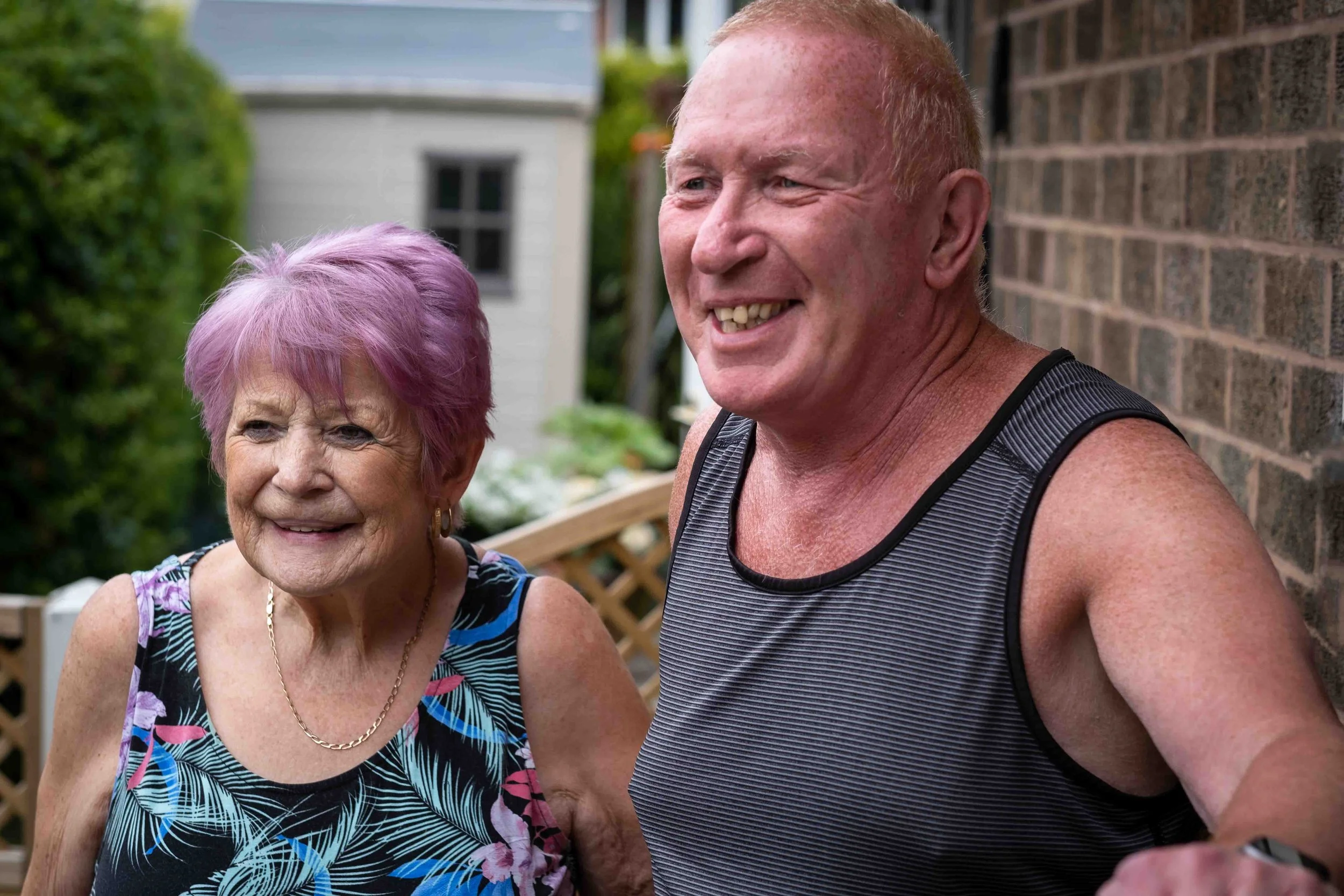 An elderly woman with purple hair smiling and a middle-aged man with reddish hair laughing outside near a brick wall.