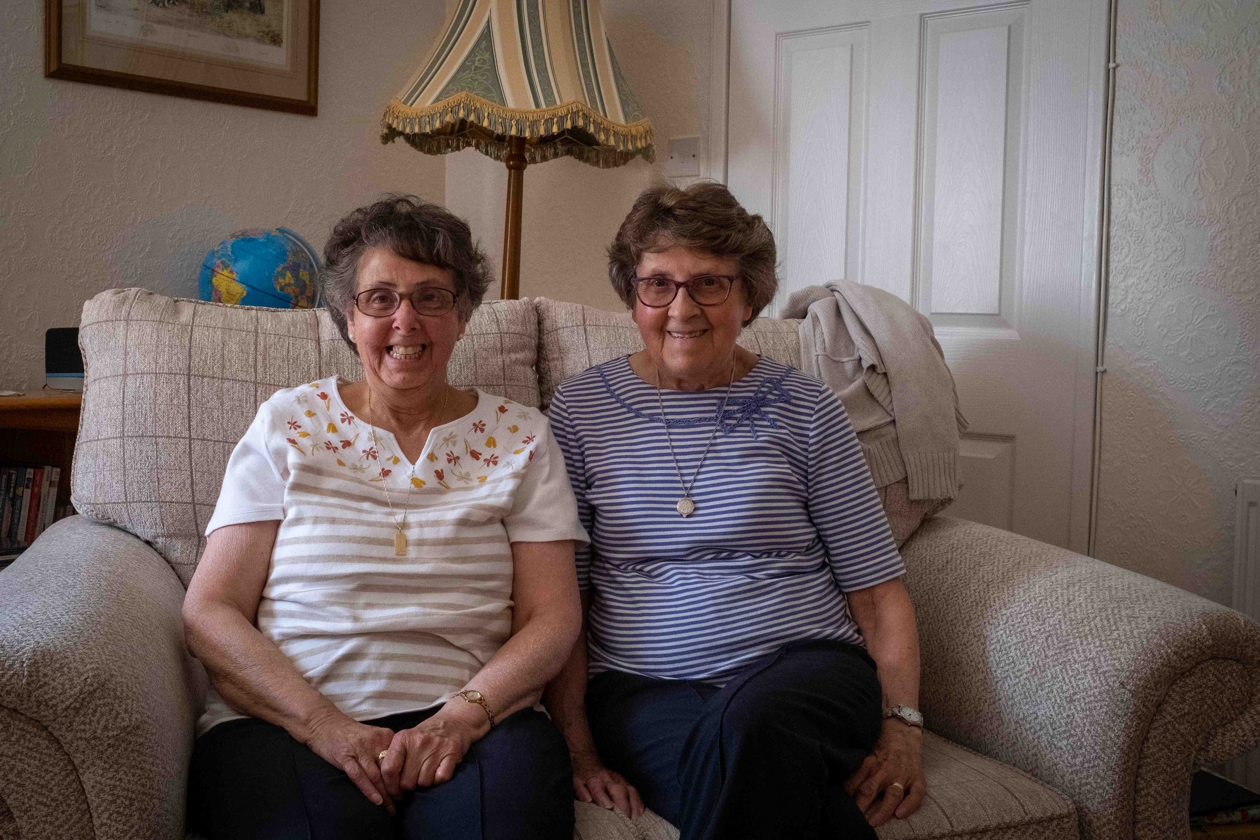 Two elderly women sitting on a beige patterned sofa in a living room, smiling at the camera. One woman is wearing glasses and a white top with floral embroidery, the other is wearing glasses and a blue and white striped top. There is a globe on a side table and a floor lamp with a fringed lampshade behind them.
