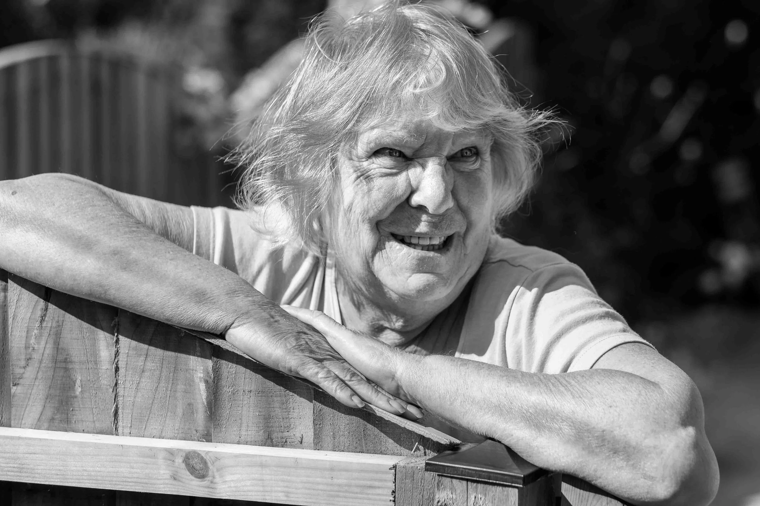 An elderly woman with light, messy hair leaning on a wooden railing outdoors, smiling and looking to her left. The image is in black and white.